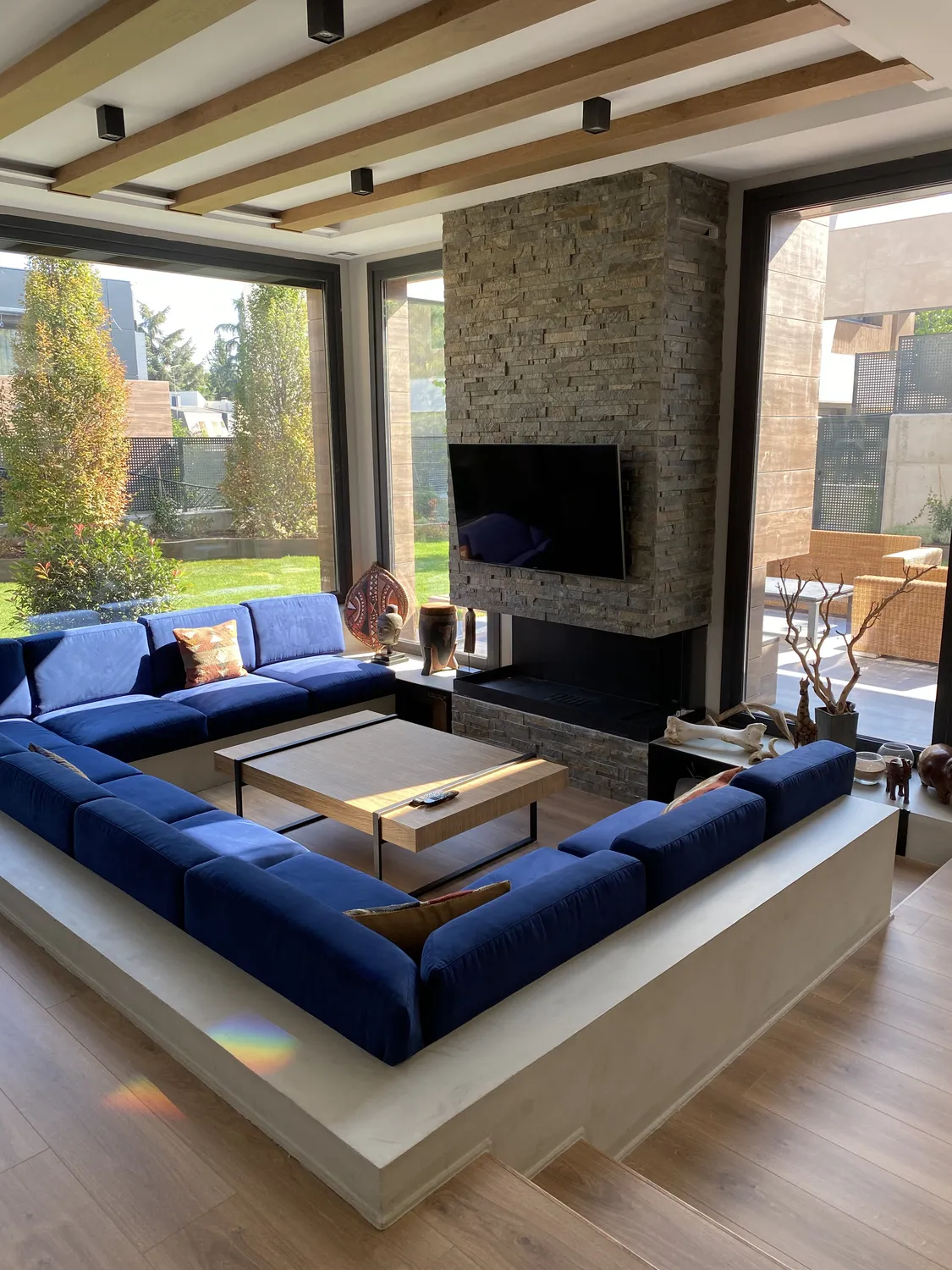 Salón con sofá encastrado azul, chimenea de piedra y grandes ventanales, vivienda en mirasierra